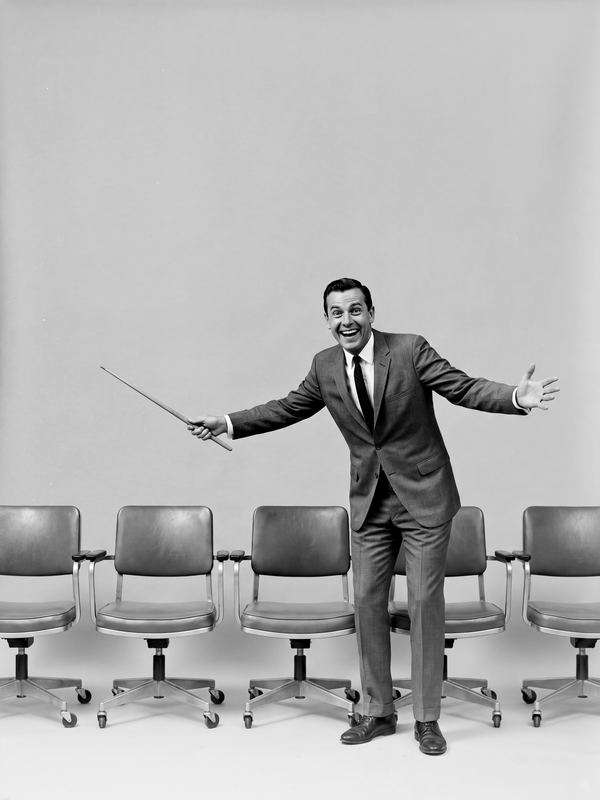 A beaming man with a pointer presenting to a row of empty office chairs. A black-and-white picture in a 1960s studio style.